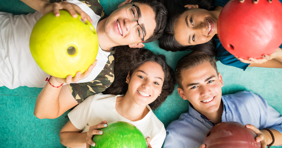 Group of 4 kids laying on the floor holding bowling balls and looking up at the camera